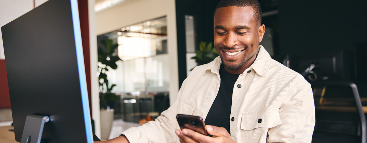 African American man smiling at his phone while he uses his pc