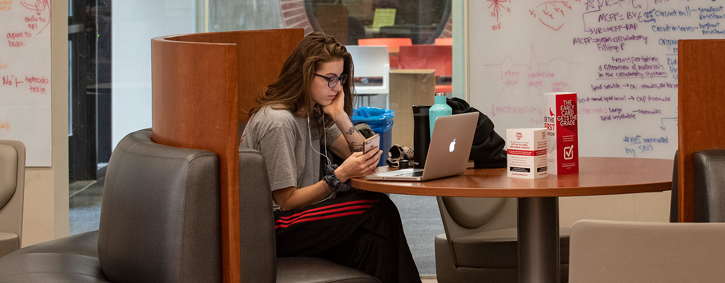 Student in library with phone and laptop.