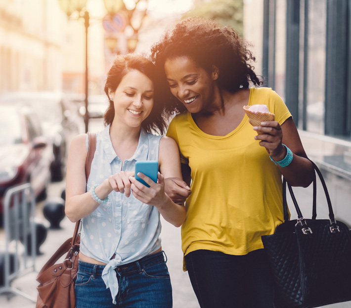 Two friends walking down the street view something on a cellphone.