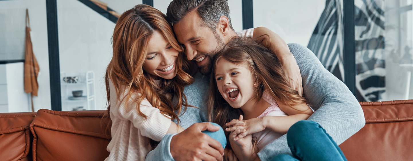 A husband and wife smile and laugh with their young daughter as they sit on the couch together.