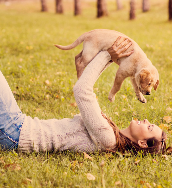 Image of a woman laying on her back in the grass, smiling at a Labrador puppy she's holding above her head