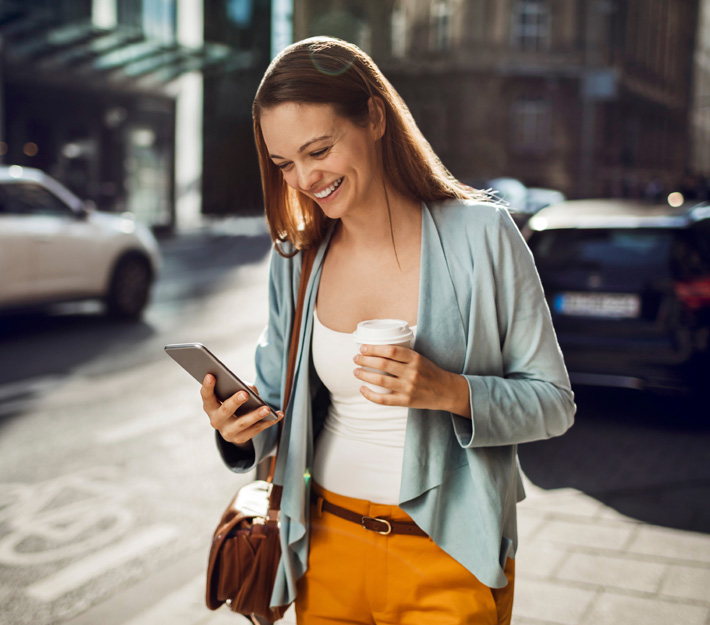 Woman smiles at phone while walking with a cup of coffee.