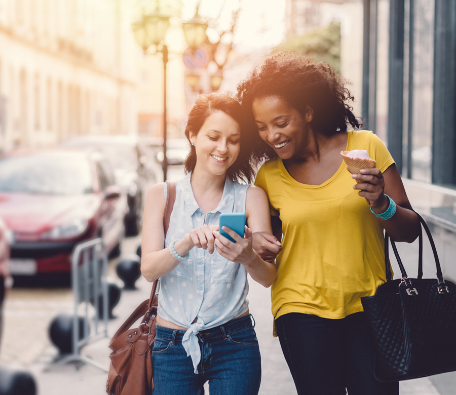 Two friends smile together while looking at a phone & walking down a street