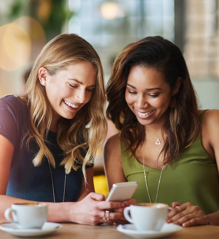 Image depicting two women sitting together at a cafe looking at a phone and smiling