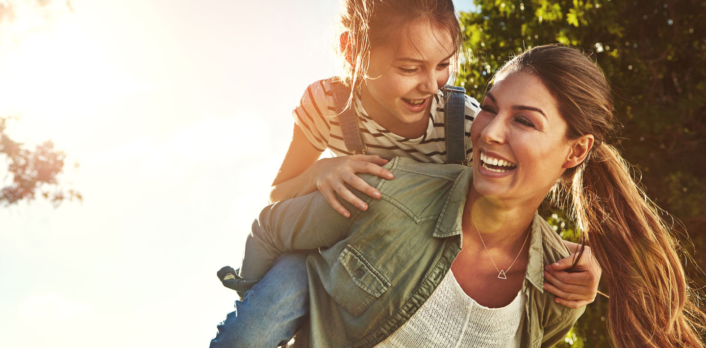 Smiling mom carries her laughing daughter piggyback.