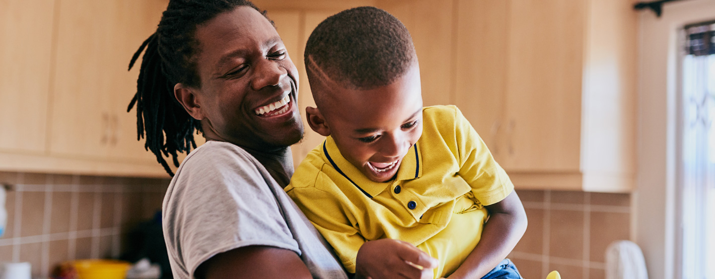 A father holds his smiling son in a kitchen.