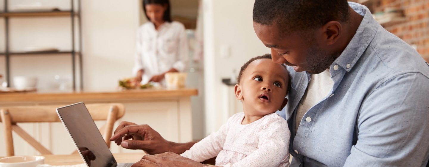 Father and his baby sit at a dining table with a laptop in front of them.