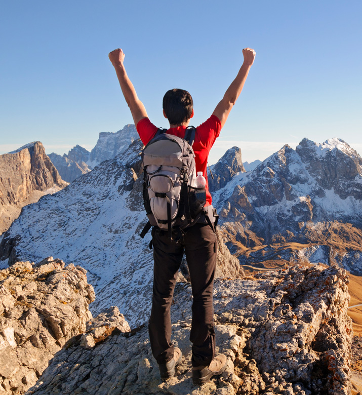 Man standing atop a mountain, hands raised in victory, with a backpack and hiking boots on.